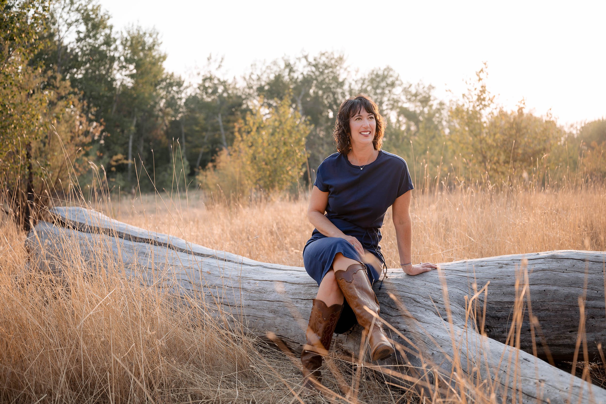 A woman in a navy blue dress and brown cowboy boots sitting on a large fallen tree trunk in a field of tall, dry grass during sunset with trees in the background.