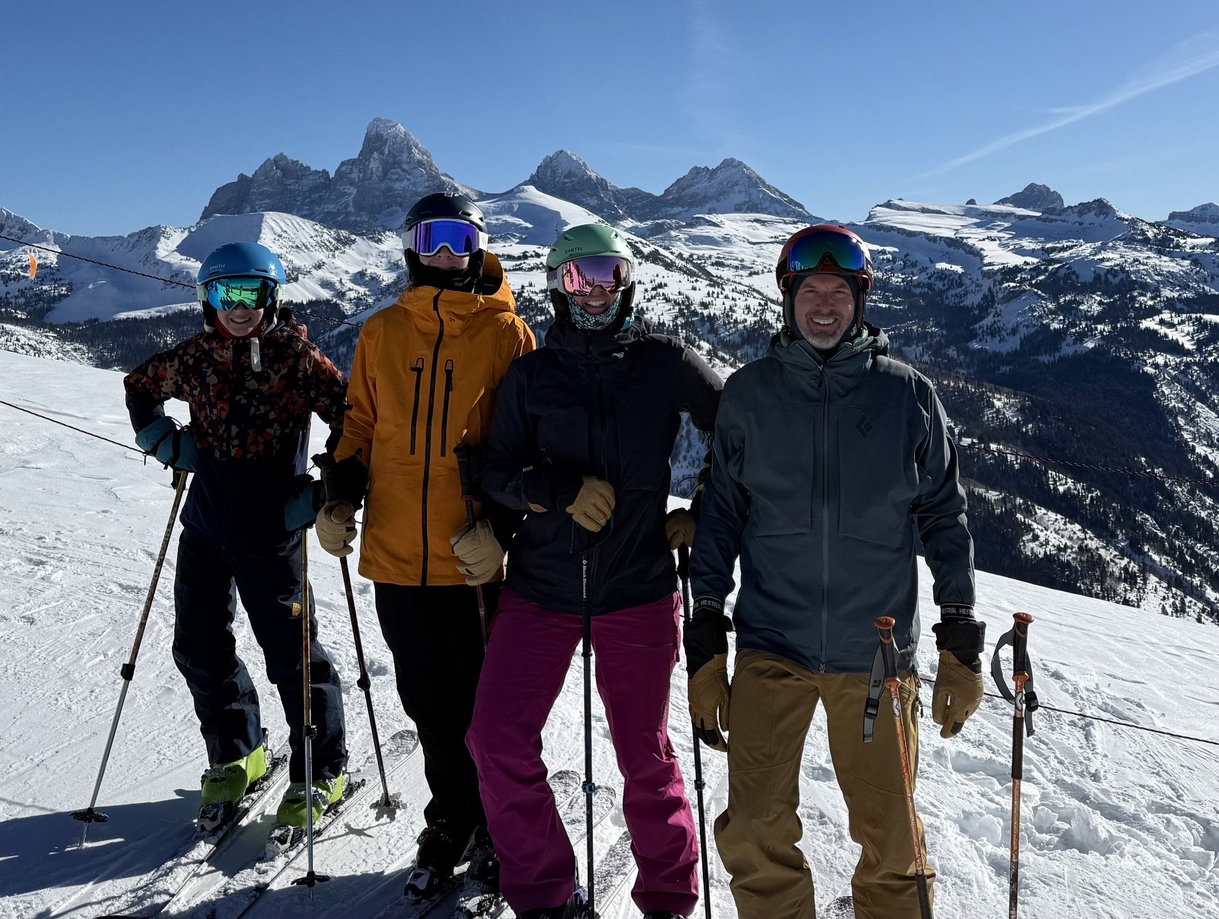 Four skiers in winter gear standing on snow with snowy mountains in the background.