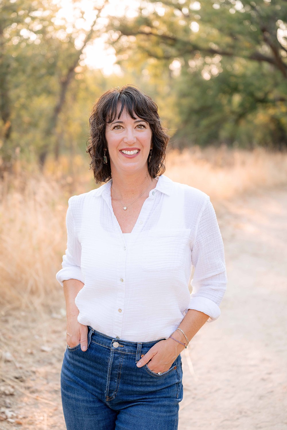 A woman with dark hair wearing a white blouse and blue jeans standing outdoors on a dirt path with trees in the background during sunset.