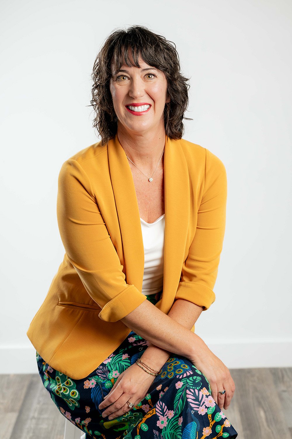 A woman with short curly dark hair wearing a yellow blazer, white top, and colorful floral pants, sitting against a plain light background, smiling.