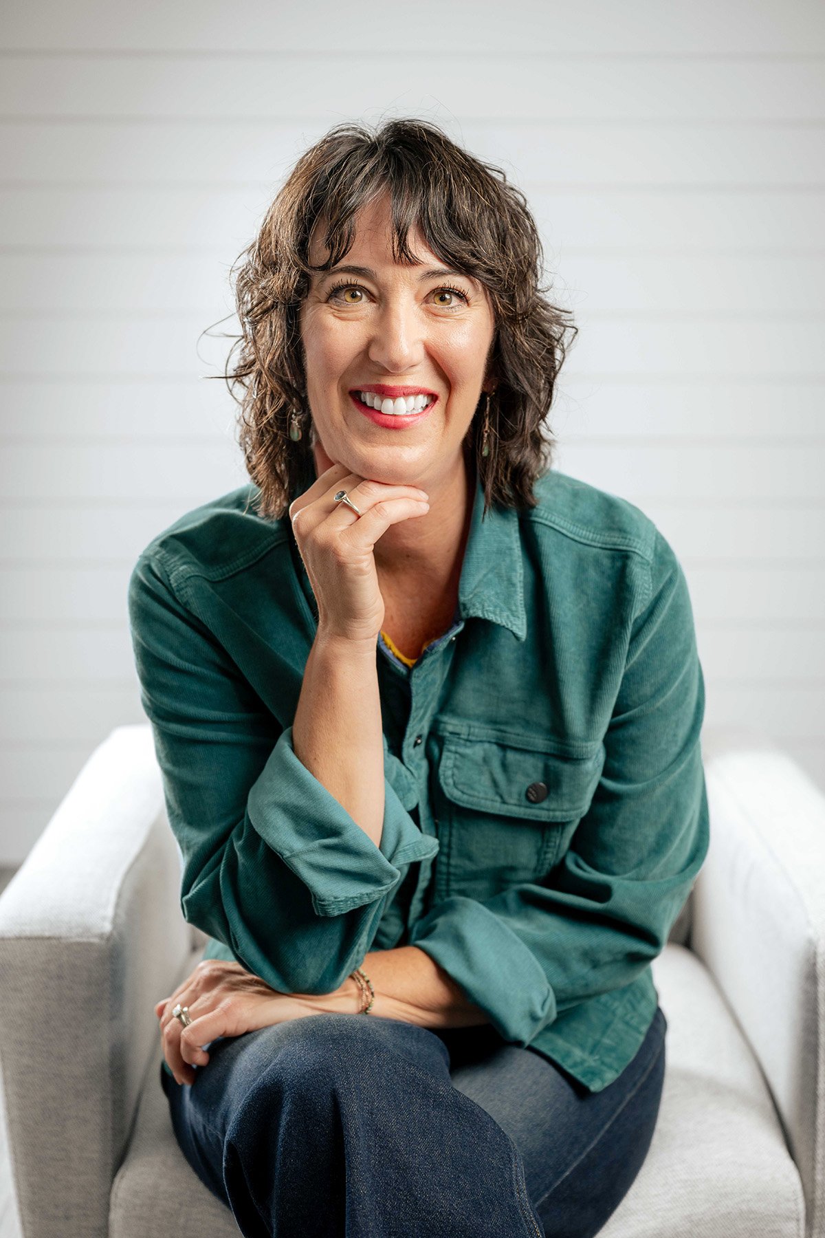 A woman with curly brown hair and hazel eyes sitting on a light gray sofa, smiling, wearing a green jacket, dark jeans, and jewelry, with a white paneled wall in the background.