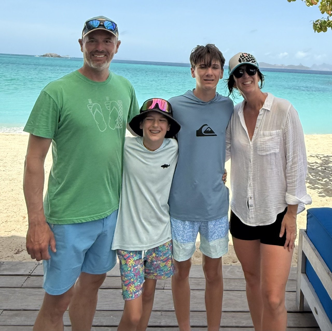 Family of four standing on a wooden deck at the beach with clear turquoise water and some islands in the background. They are smiling and dressed in casual beachwear.