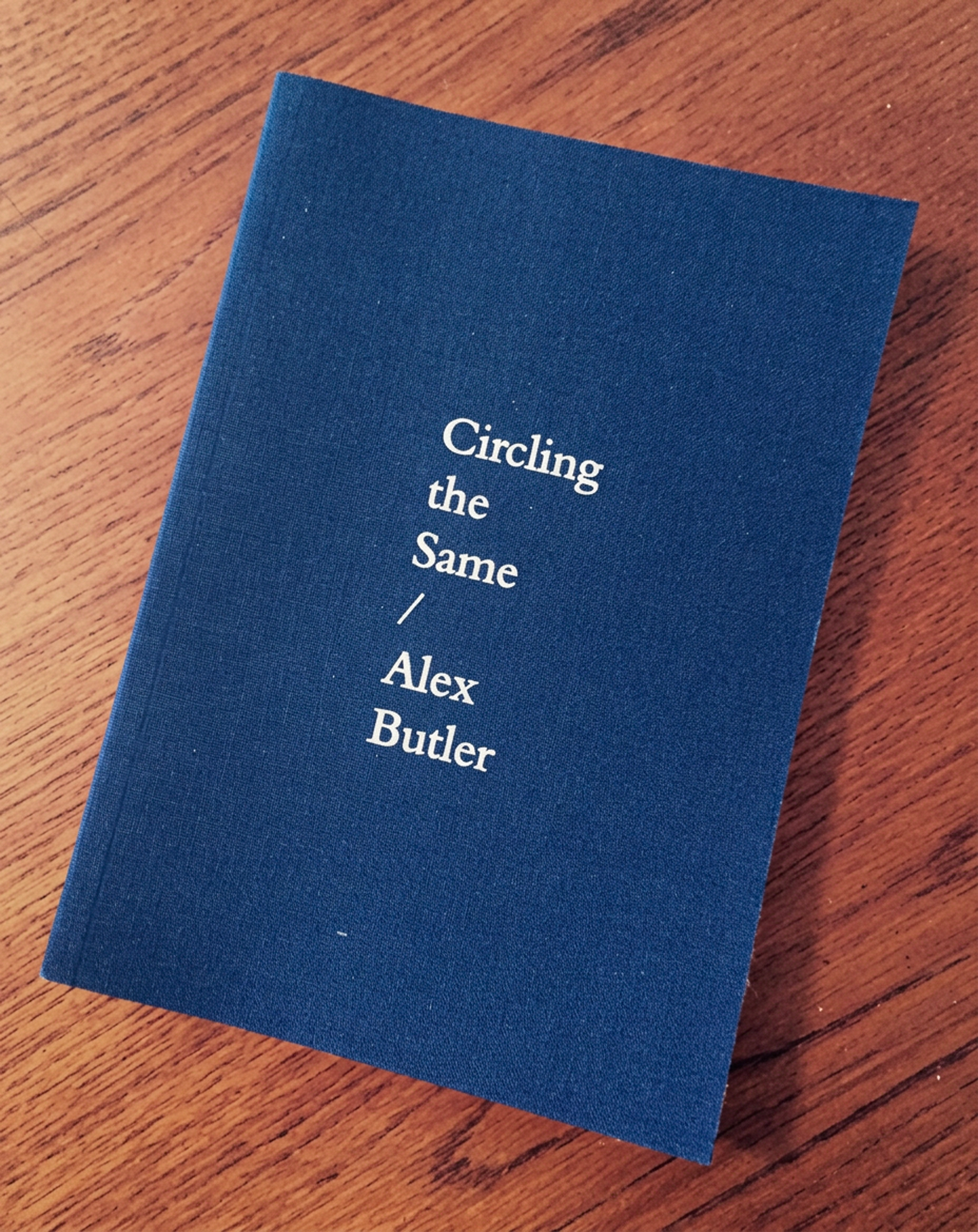 A blue book titled 'Circling the Same' by Alex Butler resting on a wooden surface.