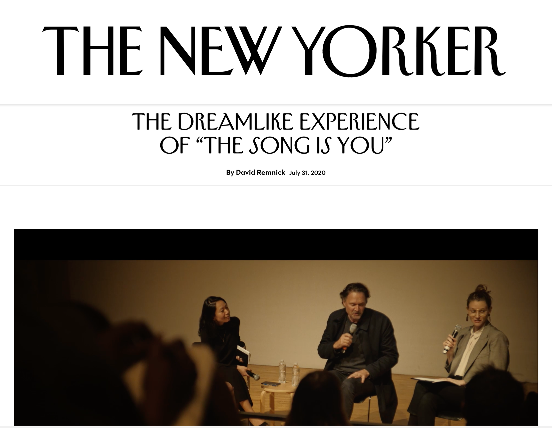 A group of people on stage at an event, one man holding a microphone and speaking, with women beside him, in front of an audience. The background features a sign that reads 'The New Yorker' and a subtitle about a 'dreamlike experience.'