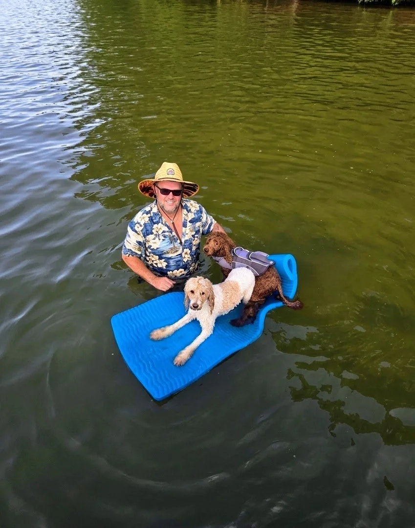 A man wearing a Hawaiian shirt, sunglasses, and a straw hat is standing in a body of water with two dogs on a blue floating mat. One dog is light-colored and laying down, while the other is dark-colored and sitting upright.