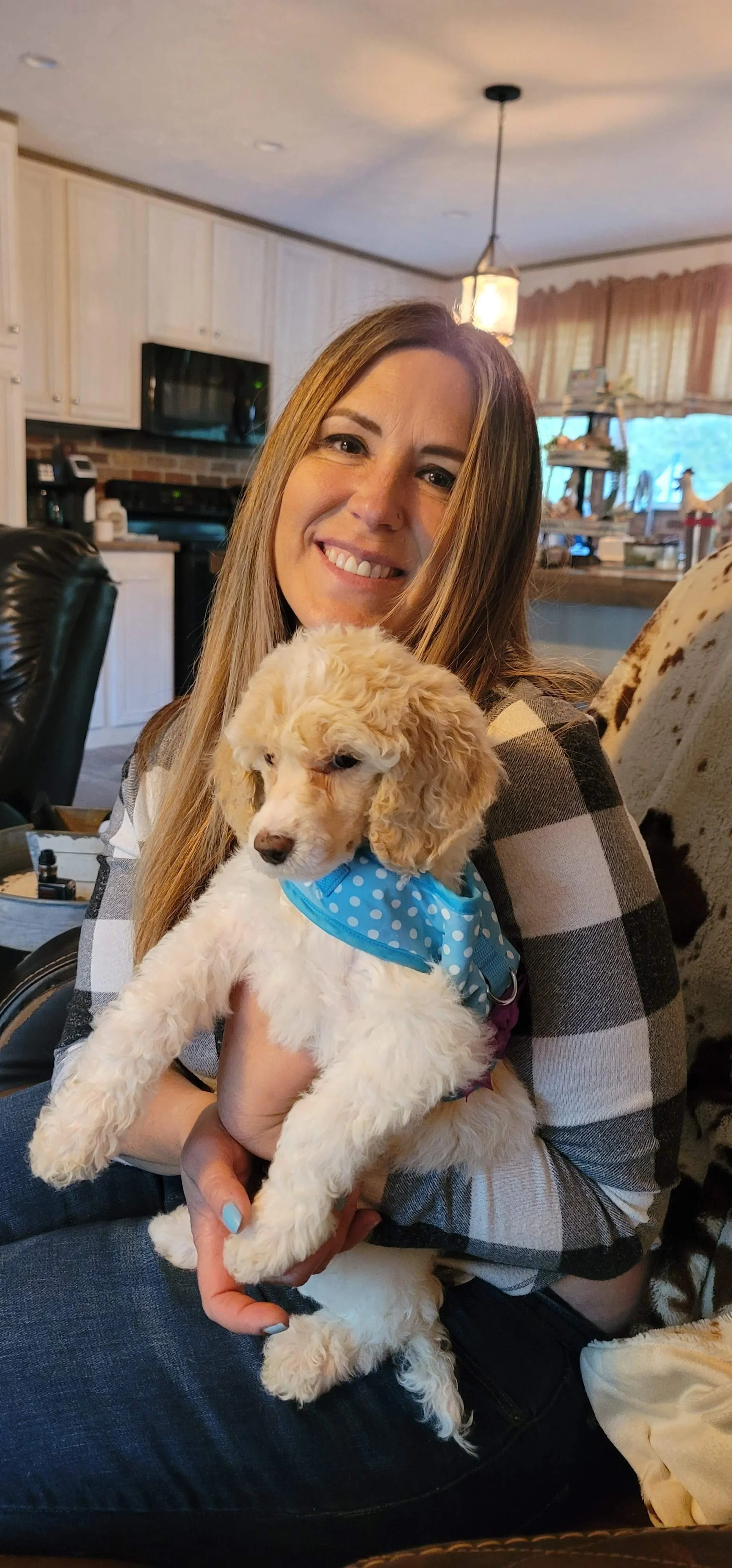 A woman with long hair smiling while holding a cream-colored poodle puppy with a blue polka dot bandana in a cozy kitchen.