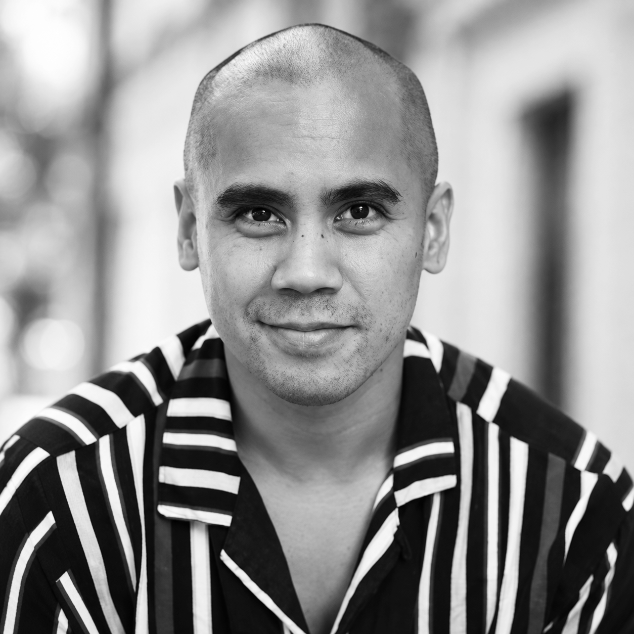 A black and white close-up portrait of a man with a shaved head, smiling, wearing a striped shirt, outdoors with blurred background.