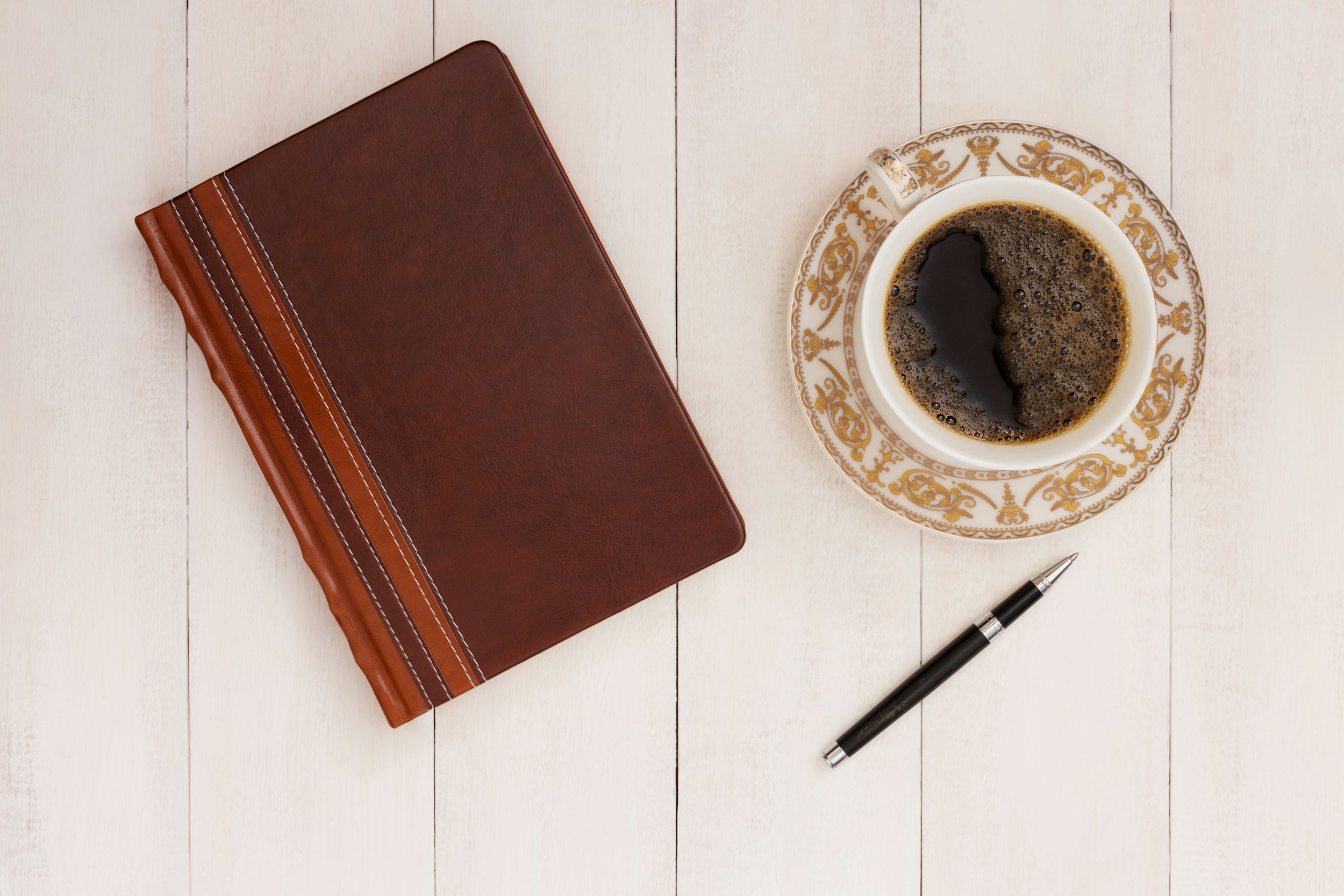 Top view of a brown leather-bound notebook, a cup of black coffee in a white cup with gold decorative rim and handle, placed on a matching saucer, a black pen with silver accents, on a white wooden surface.