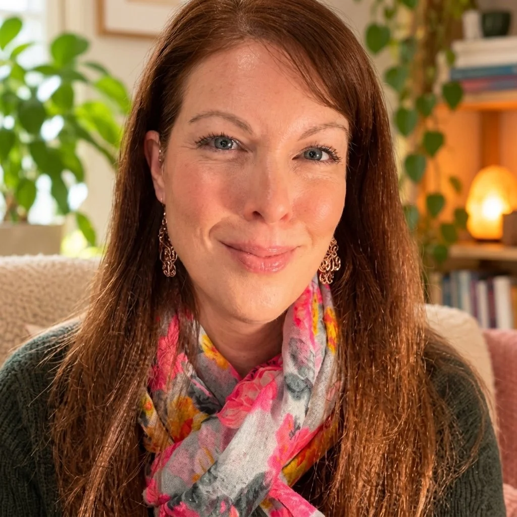 A smiling woman with long brown hair, wearing earrings and a floral scarf, sitting in a cozy, well-lit room with plants and books in the background.