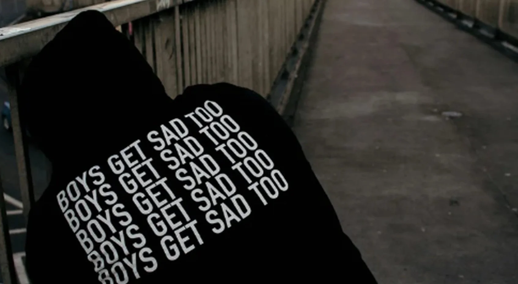 Person sitting on a sidewalk wearing a black hoodie with white text that says 'BOYS GET SAD TOO' repeated in lines.