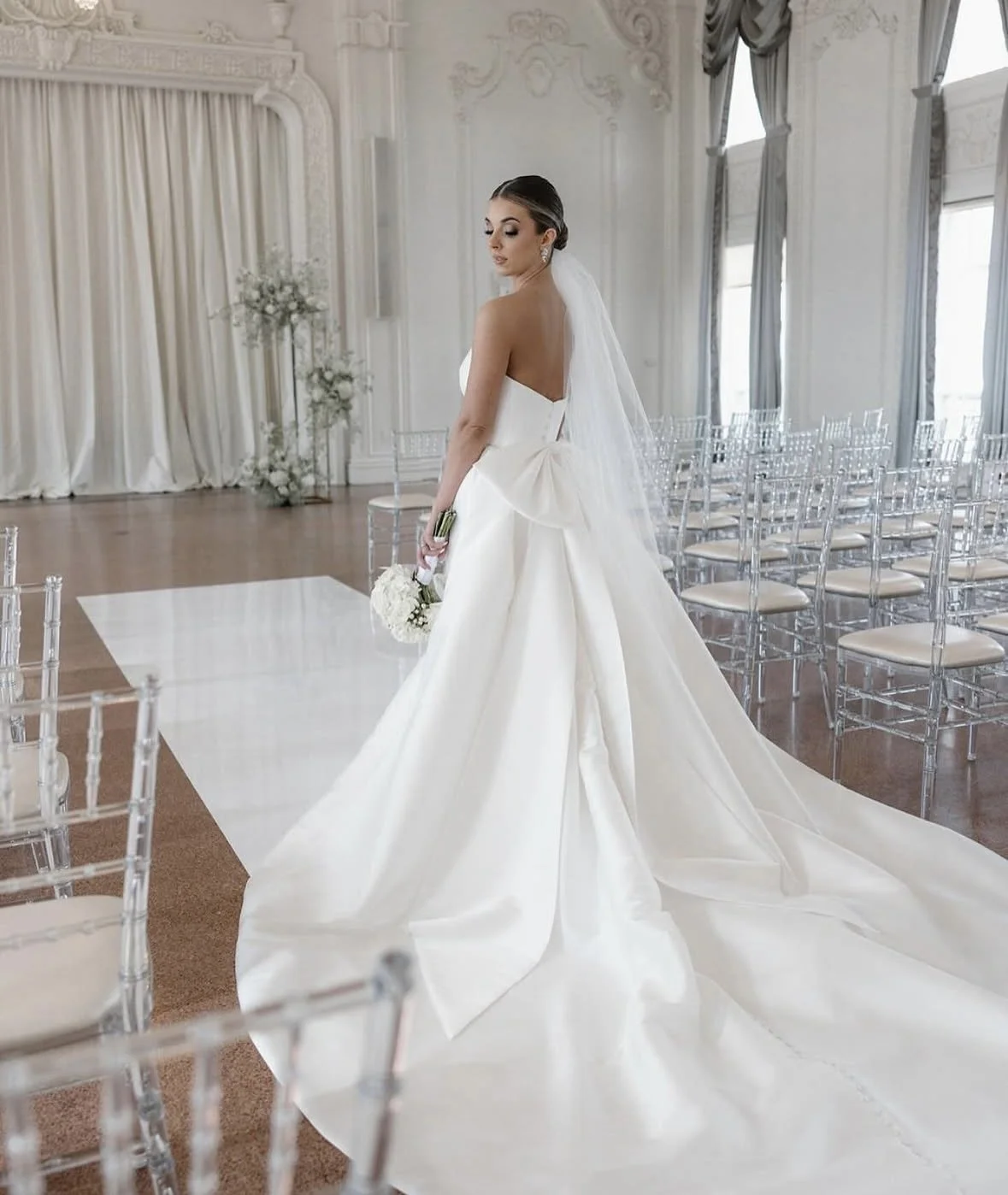 A bride in a white wedding gown holding a bouquet standing in an elegant, ornate wedding hall with rows of transparent chairs and floral decorations.