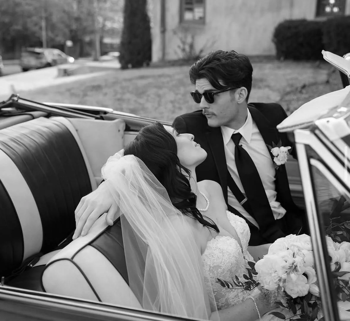 A black and white photograph of a bride and groom sitting in a vintage convertible car. The bride is leaning back with her eyes closed, wearing a veil and holding a bouquet of flowers. The groom is looking at her, wearing sunglasses and a suit with a boutonniere. They are outdoors near a grassy area and houses.