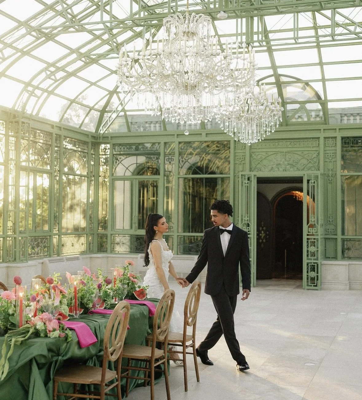 A couple dressed in formal wedding attire, holding hands, walking inside a conservatory with green ornate metal and glass architecture, large crystal chandeliers, and a decorated dining table with pink flowers and candles.
