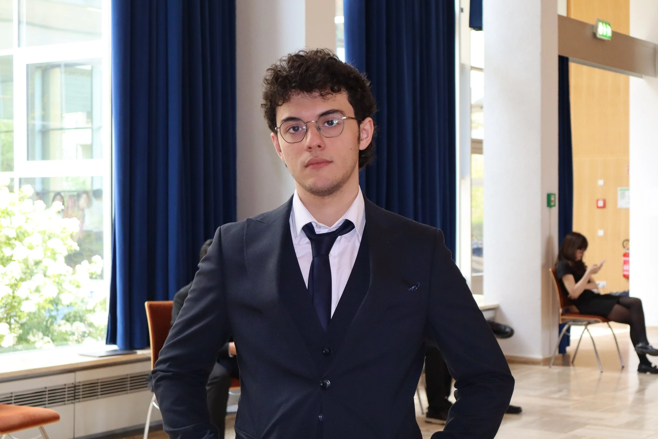 A young man with dark curly hair, glasses, wearing a black suit and tie, sitting indoors with a serious expression. Background includes blue curtains, large windows, and a woman seated in the distance.