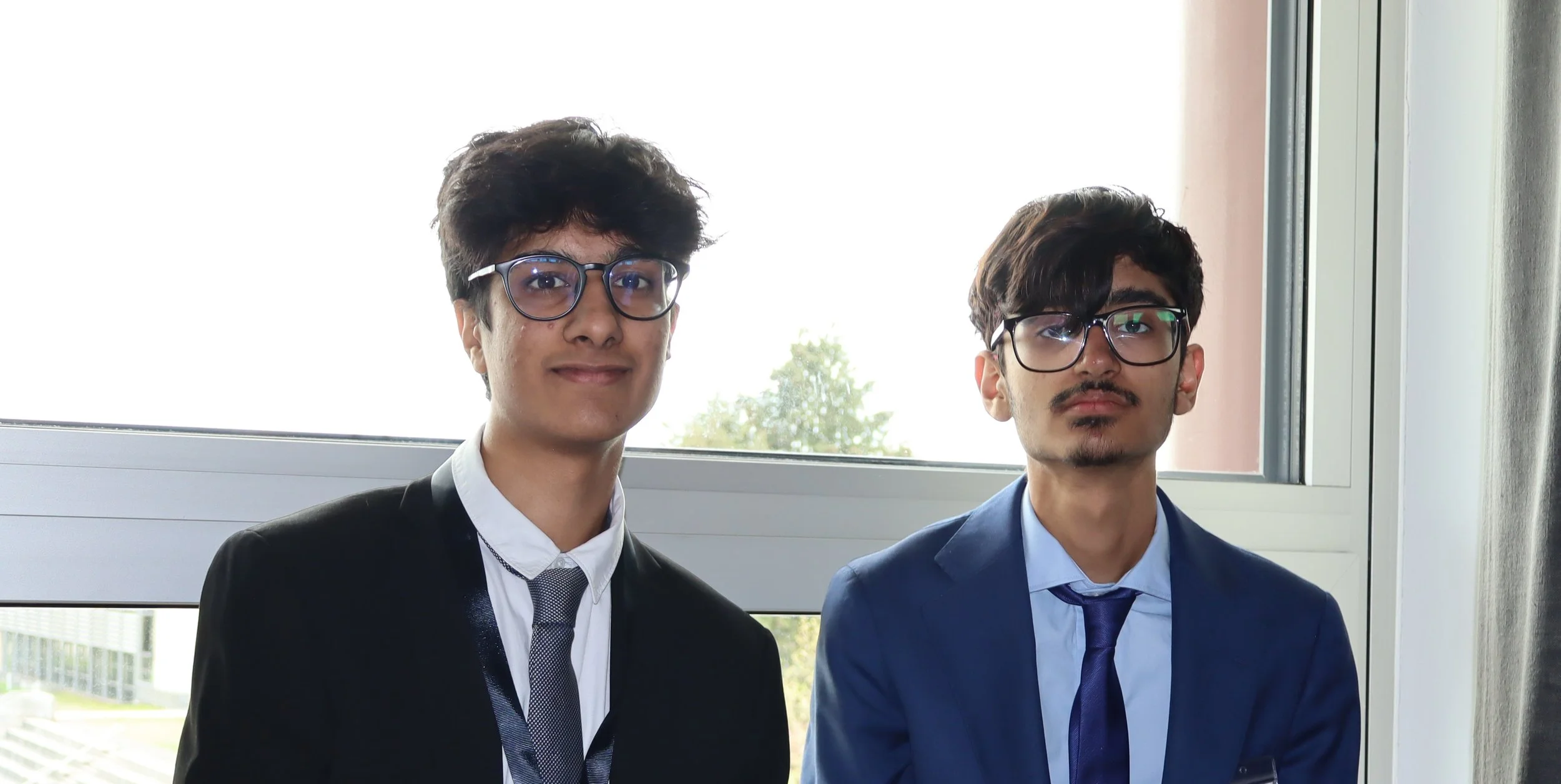 Two young men dressed in suits and glasses standing in front of a large window with trees visible outside.