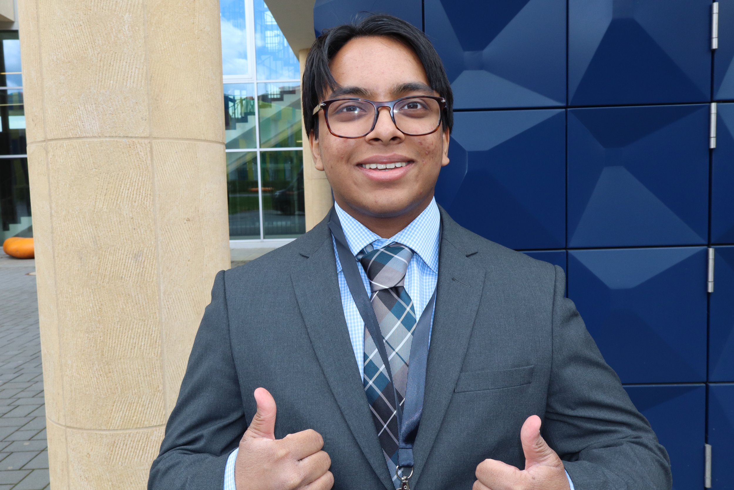 A young man wearing glasses and a suit, smiling and giving an affirmative thumbs-up gesture with both hands, standing outside a modern building with large windows and geometric blue wall panels.