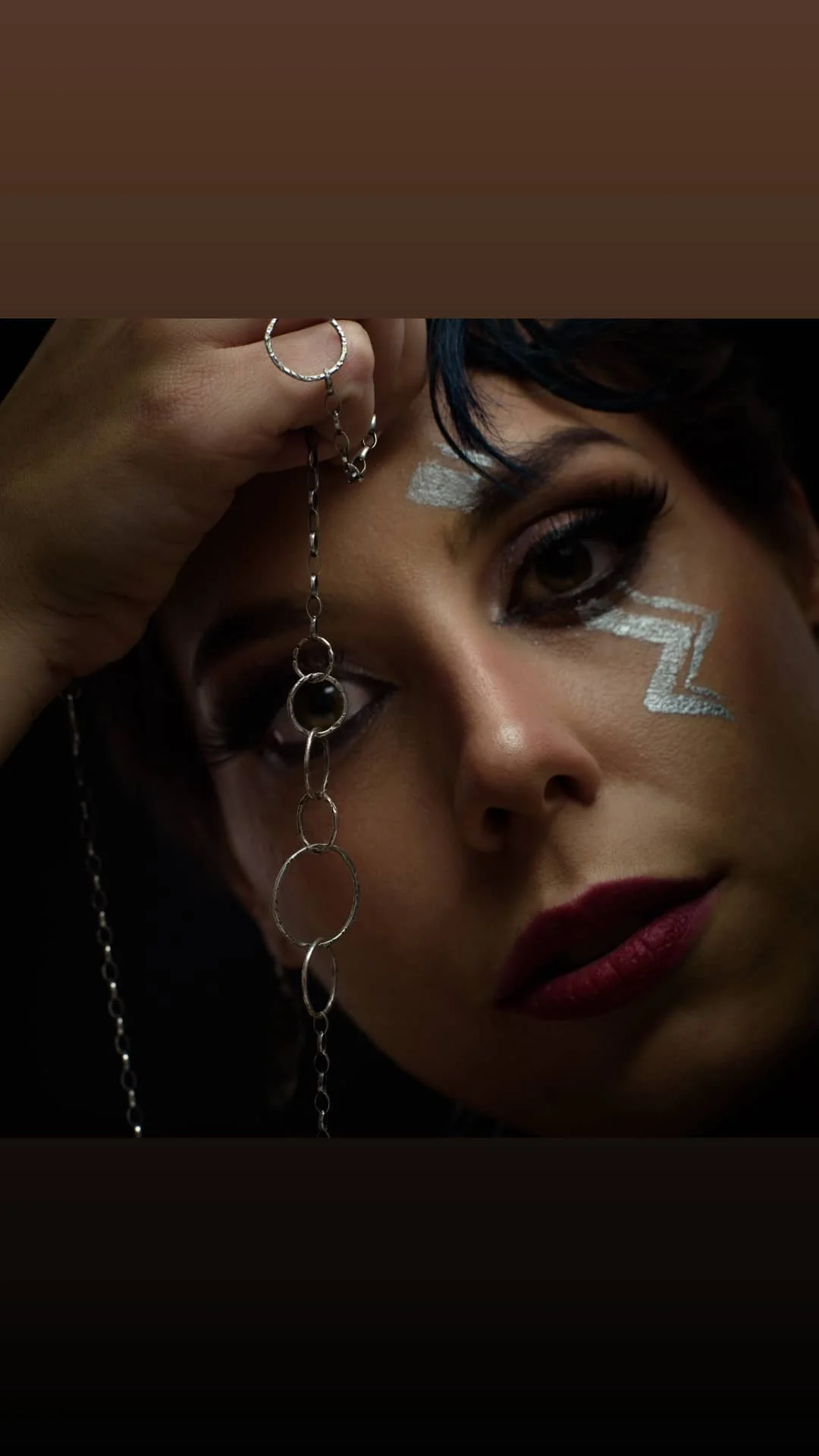 A close-up portrait of a person with bold makeup, jewelry, and a unique metallic face paint design.