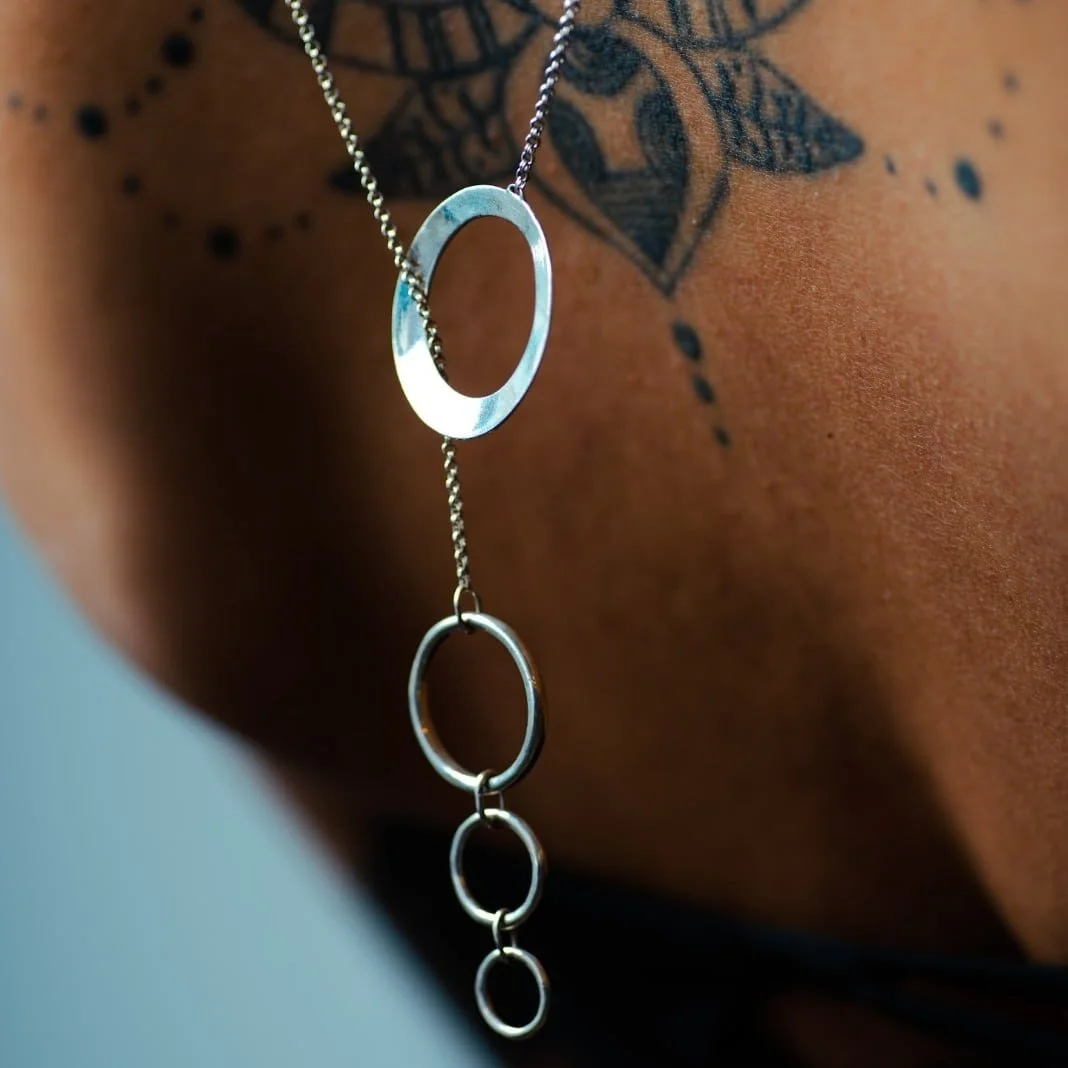 Close-up of a silver necklace with circular links worn on a person's neck, revealing part of a tattoo on the skin.