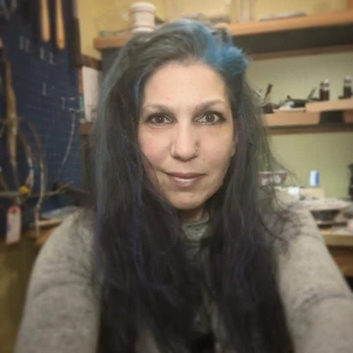 A woman with long dark hair and a small blue streak, smiling in a workshop setting with shelves and tools in the background.