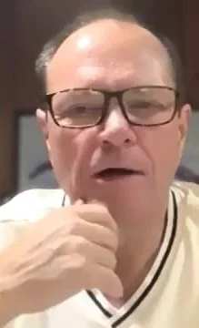 Close-up of a man with glasses, short hair, wearing a white shirt with a black stripe, speaking or thinking with his hand near his chin.