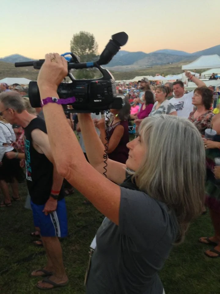 A woman with gray hair filming at an outdoor event during sunset with a video camera, surrounded by a crowd of people, tents, and mountains in the background.