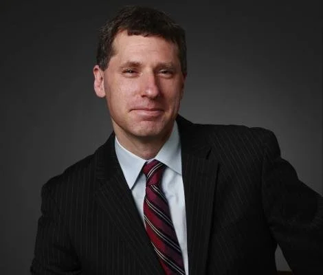 A man in a dark pinstripe suit with a white shirt and a striped maroon tie, posing against a dark gray background.