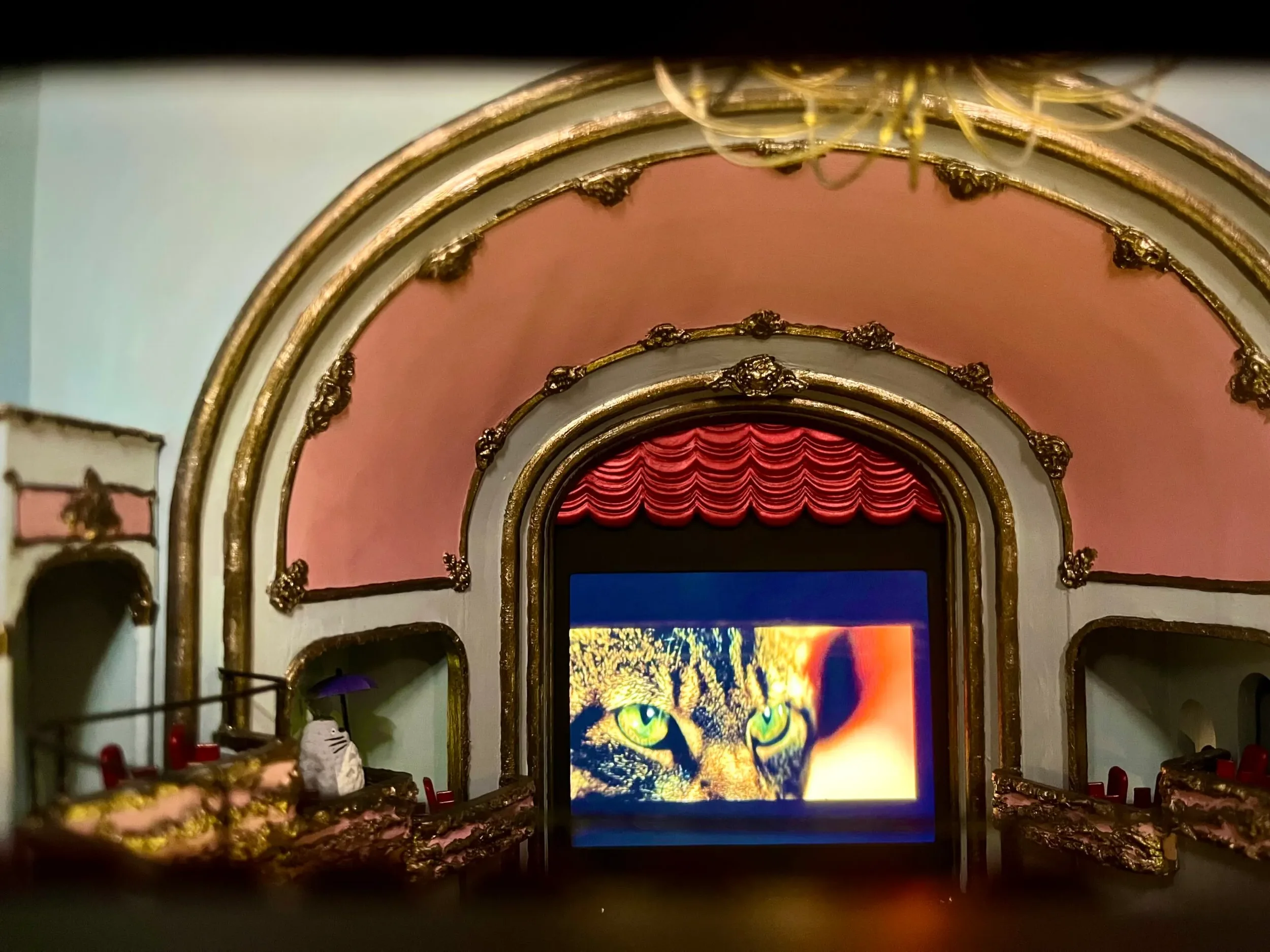 The image shows the interior of a theatrical stage with ornate gold trim and pink walls. There is a large, colorful digital display at the back featuring a cat's face with bright green eyes, and part of a red curtain above it.