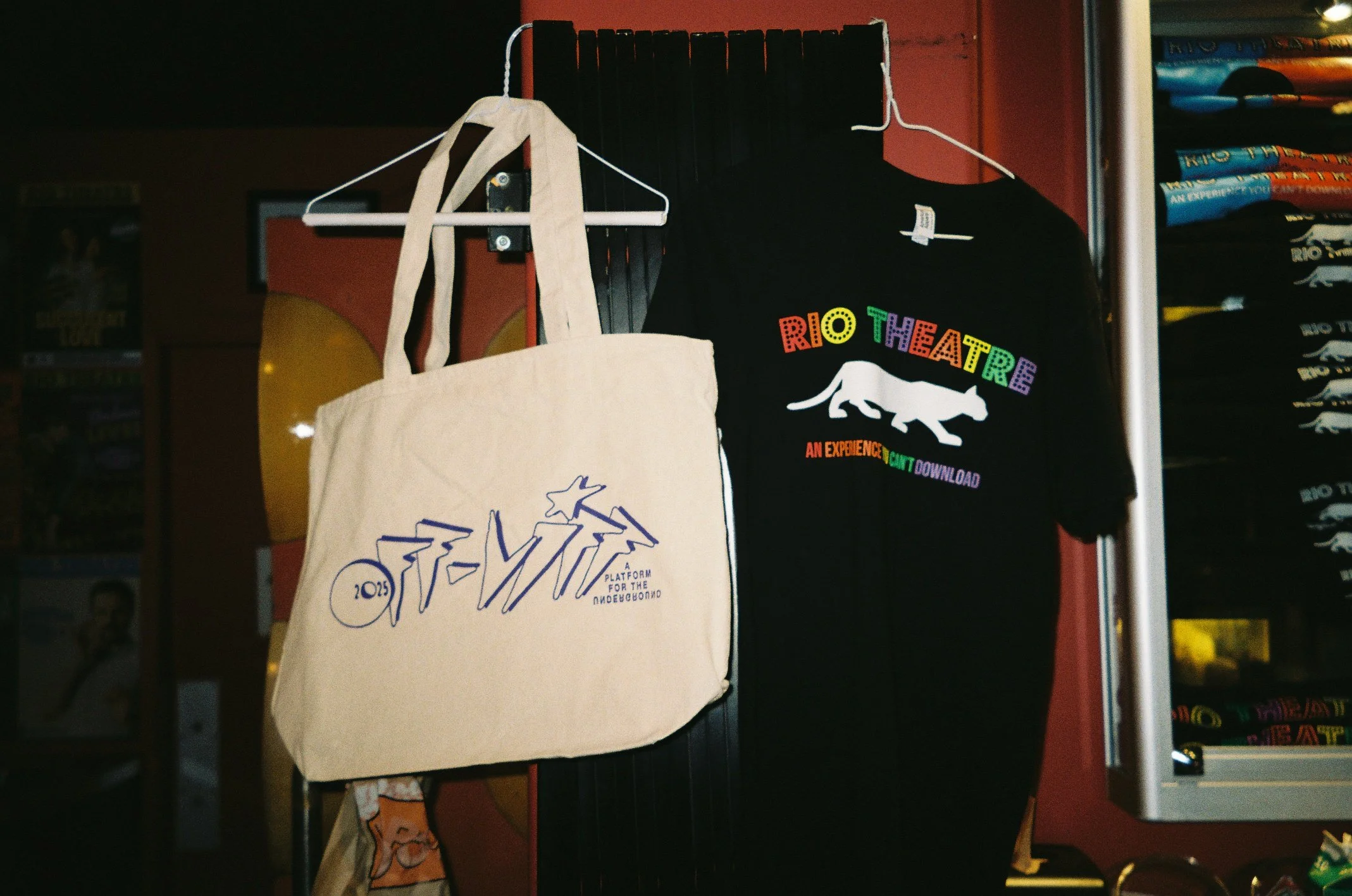 Various merchandise items, including a tote bag with a graphic design and a T-shirt with rainbow-colored text and a white animal graphic, hanging on hooks in a retail store.