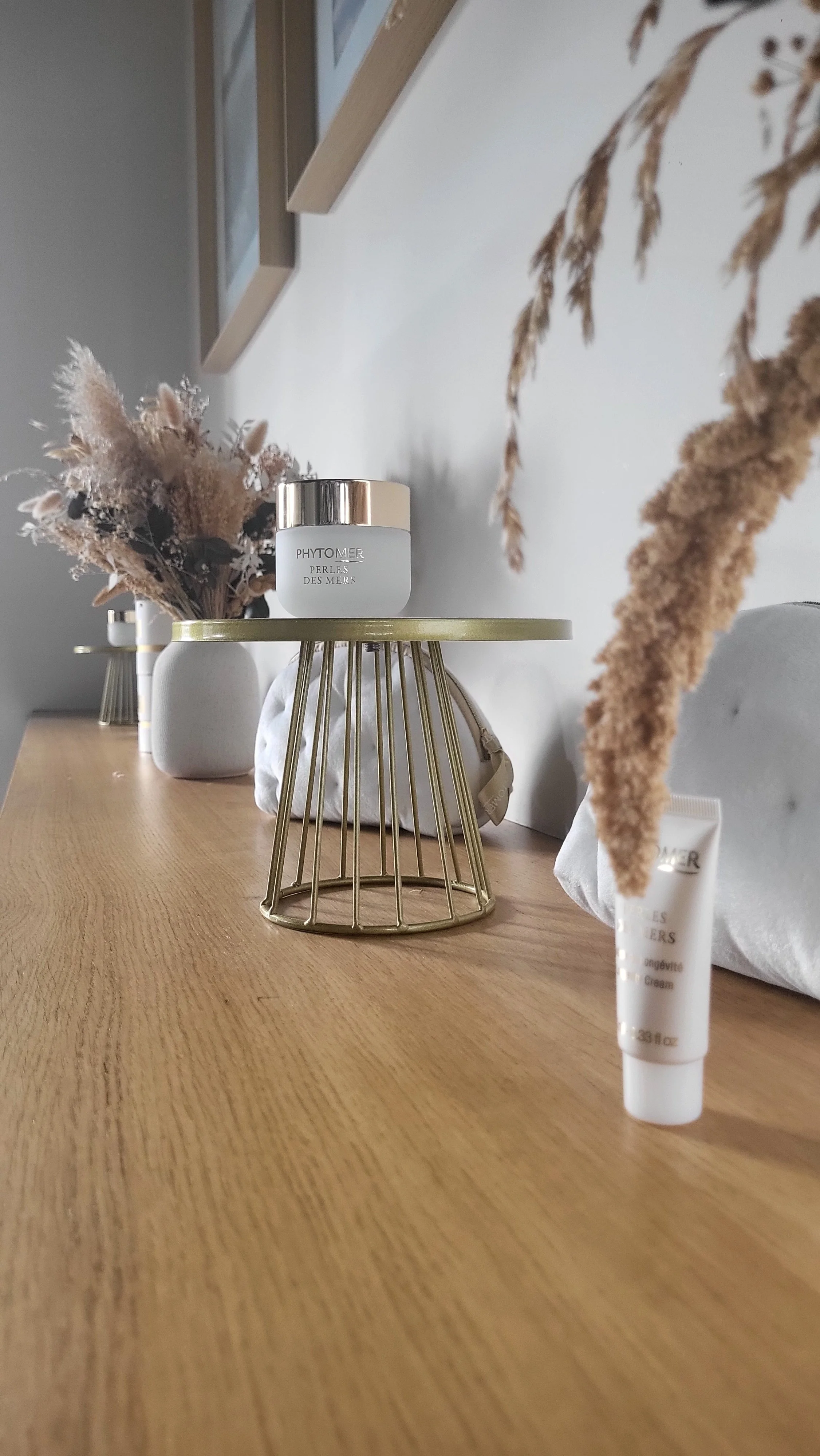 Une table en bois clair avec diverses astuces de soins et des décorations, dont une vase avec des fleurs séchées, un pot en céramique blanche, et plusieurs contenants de produits de beauté, dans un intérieur moderne et minimaliste.