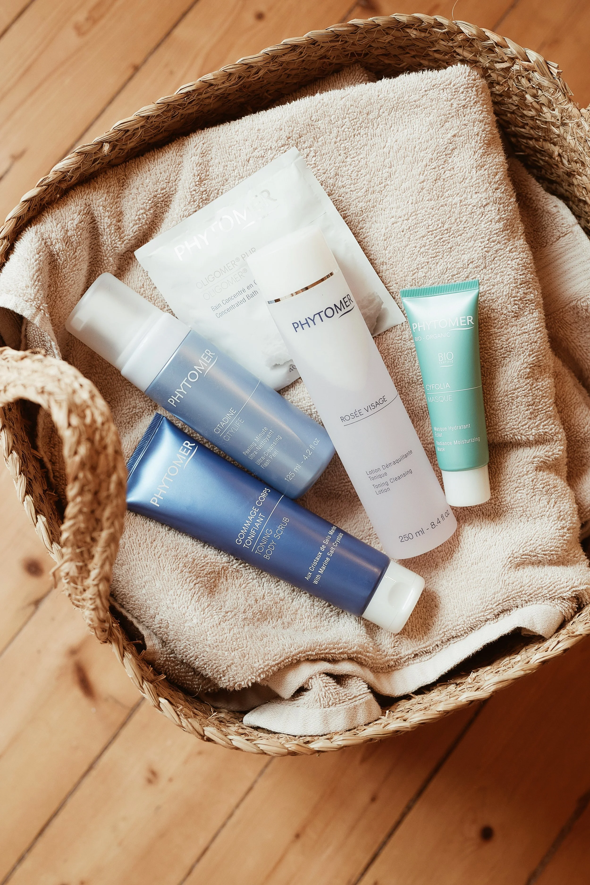 Un panier en osier contenant des produits de soins de la marque Phytomer, posés sur une serviette en tissu, sur un sol en bois.