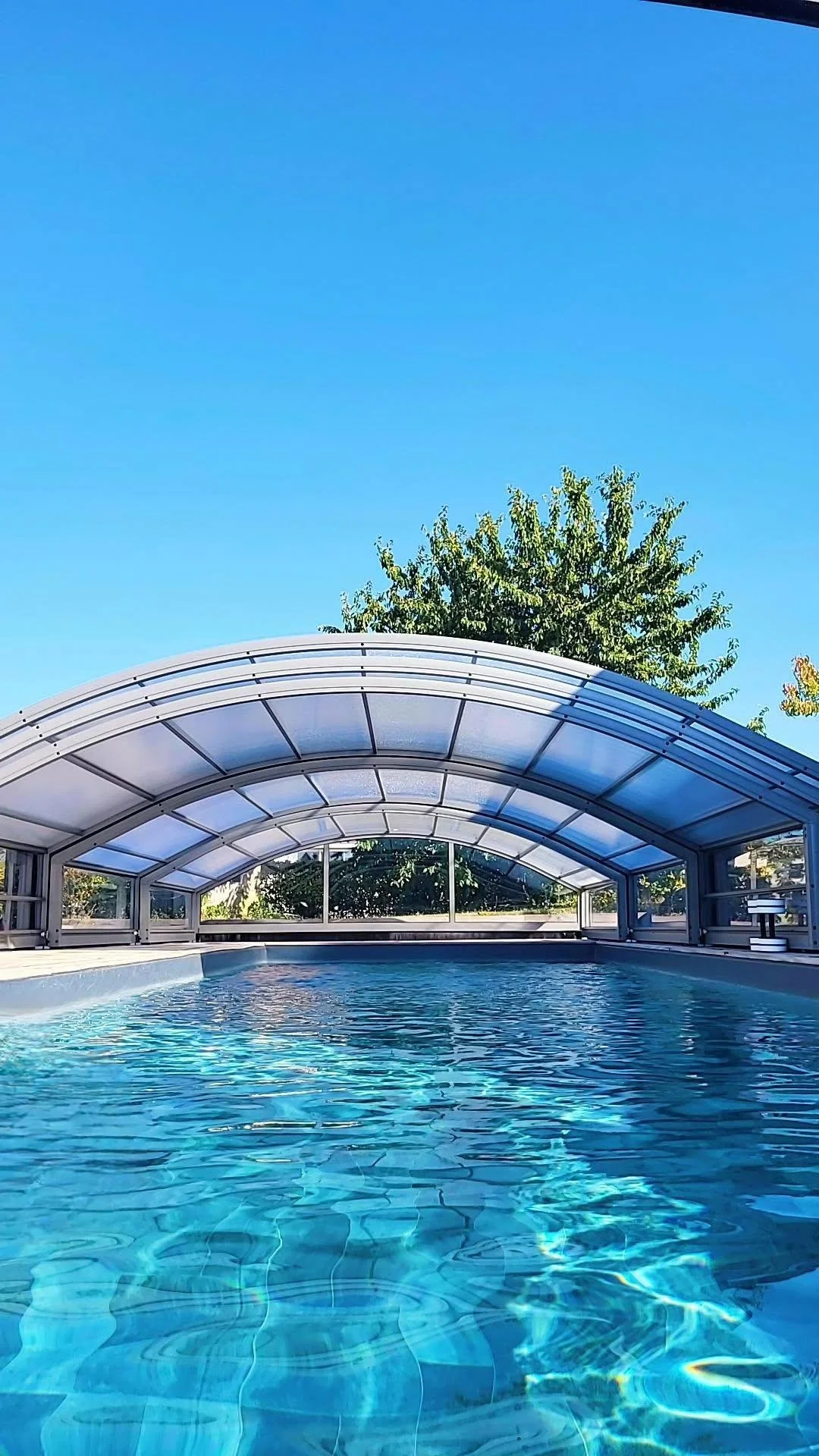 Piscine à débordement couverte, avec un toit arqué en plexiglas, entourée d'arbres sous un ciel bleu.