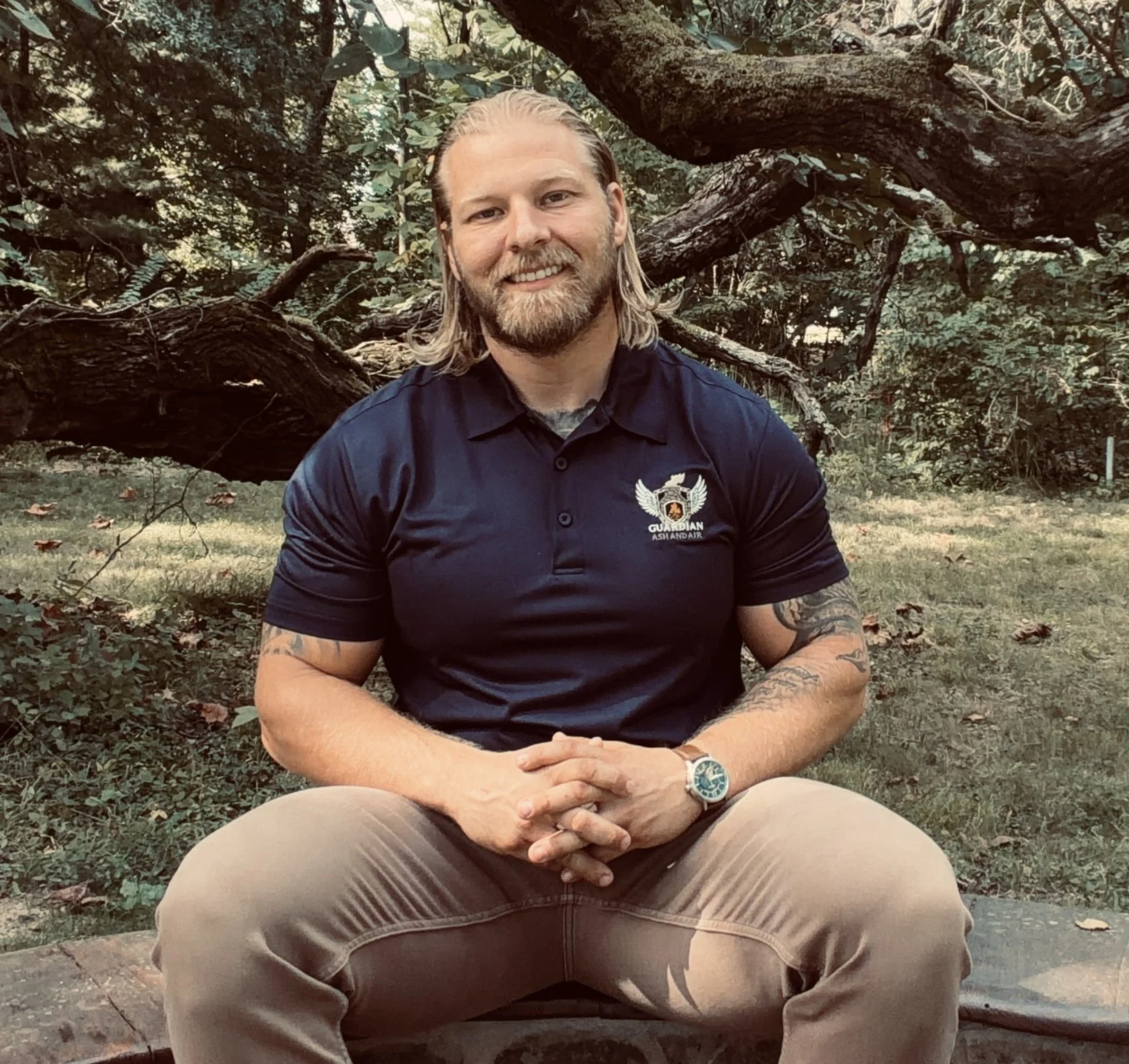 A man with long blonde hair and a beard, smiling, sitting outdoors in a wooded area with a large fallen tree in the background. He is wearing a black polo shirt with a logo and beige pants.