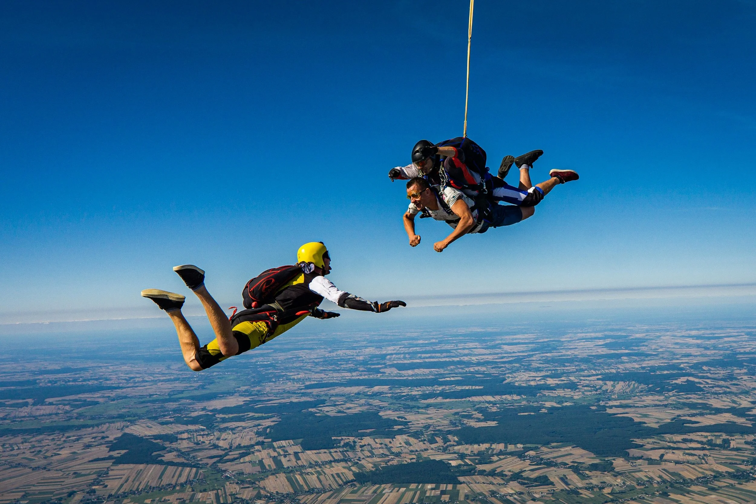 Zwei Fallschirmspringer fliegen während des freien Falls über einer landwirtschaftlichen Landschaft mit Feldern und Flüssen, während ein dritter Springer den Flug beobachtet.