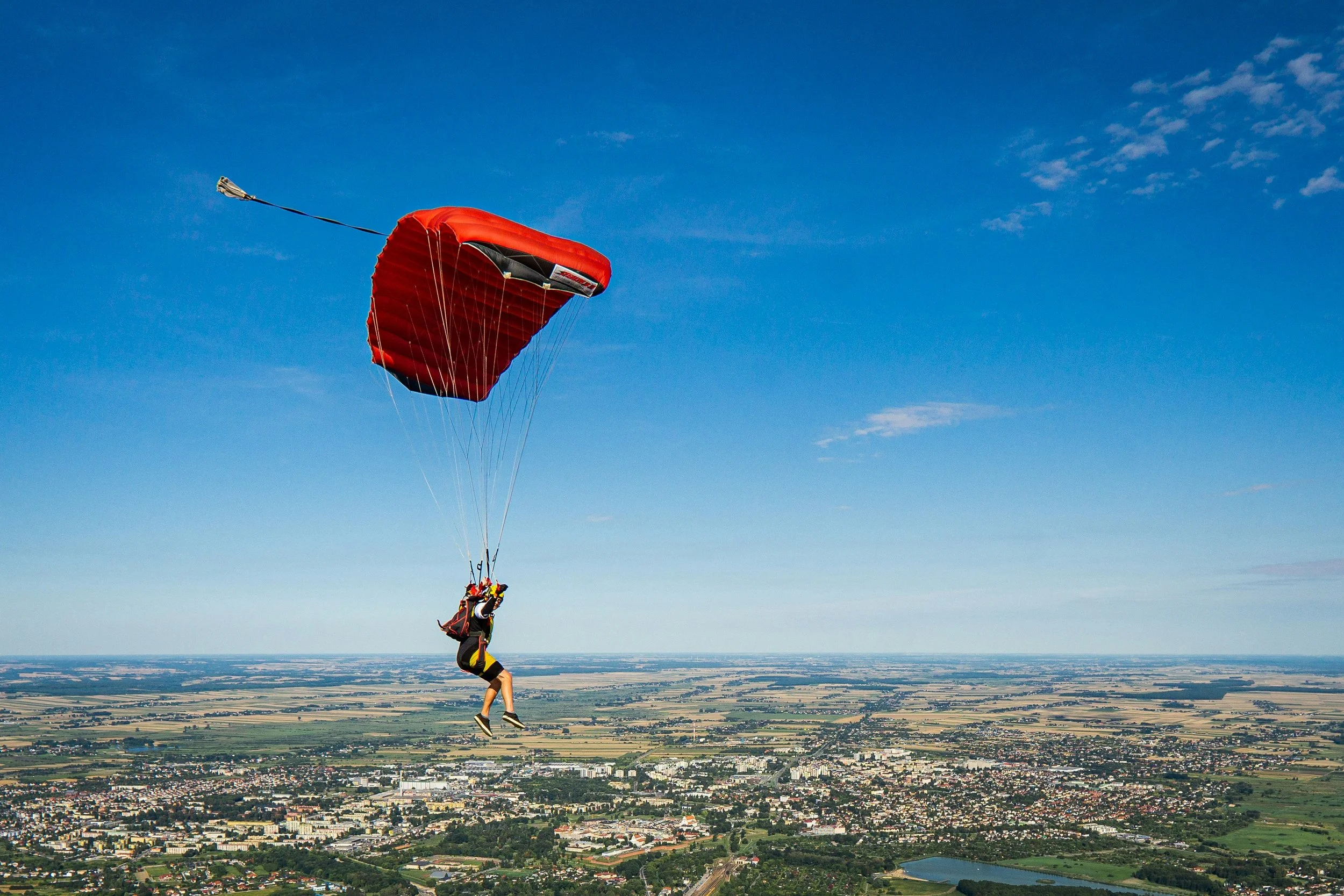 Eine Person mit Fallschirm in der Luft über einer Stadt und Landschaften darunter, blauer Himmel mit wenigen Wolken.