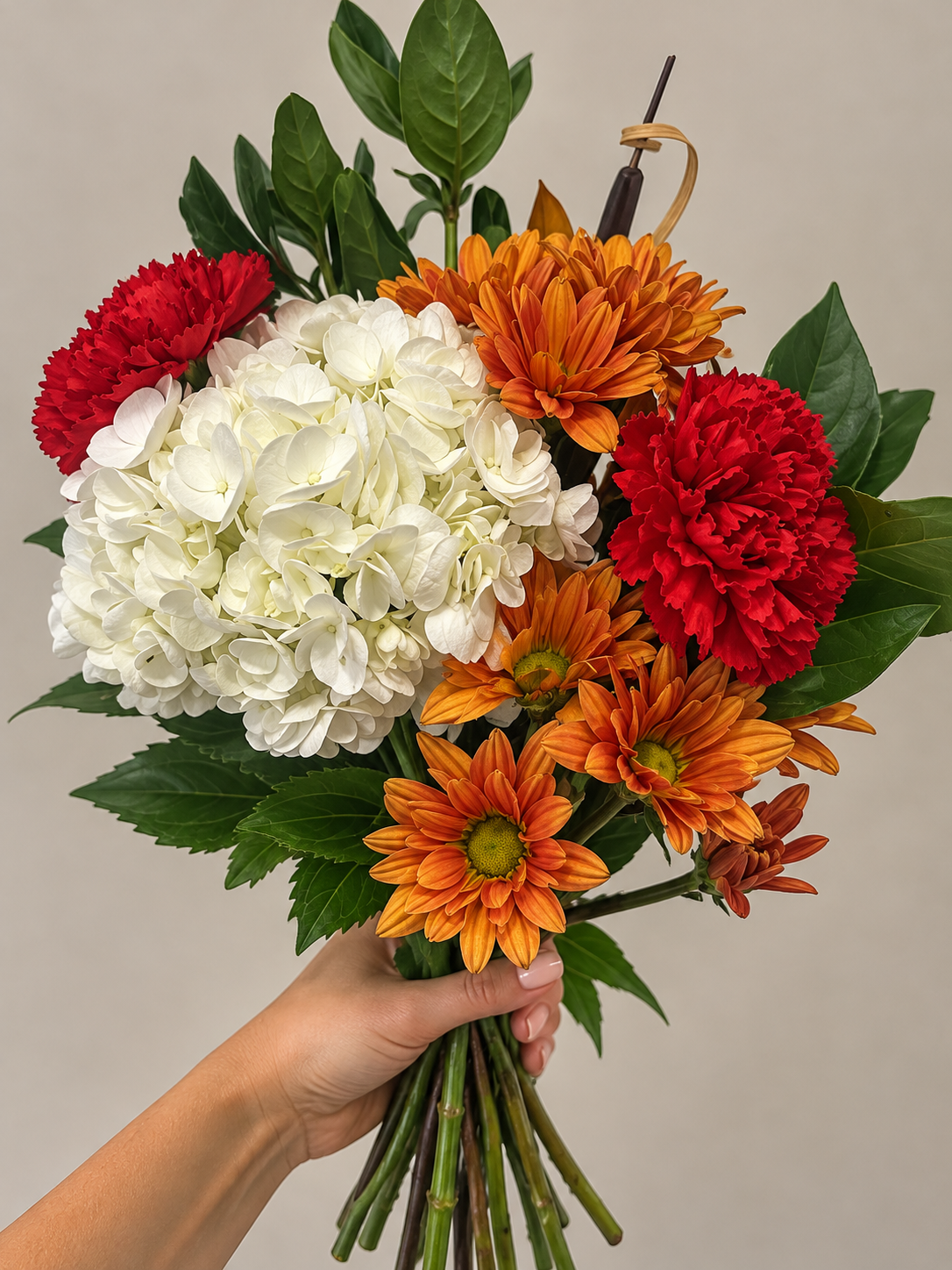 Hand holding a colorful bouquet of white hydrangeas, orange daisies, and red carnations with green leaves.