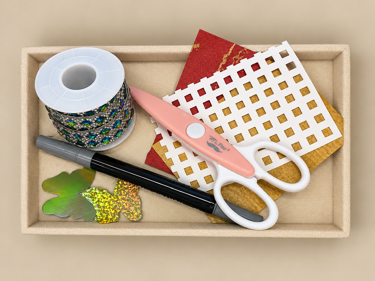 A beige craft organizer tray containing rhinestone jewelry, scissors, decorative paper, a marker, and leaf embellishments.