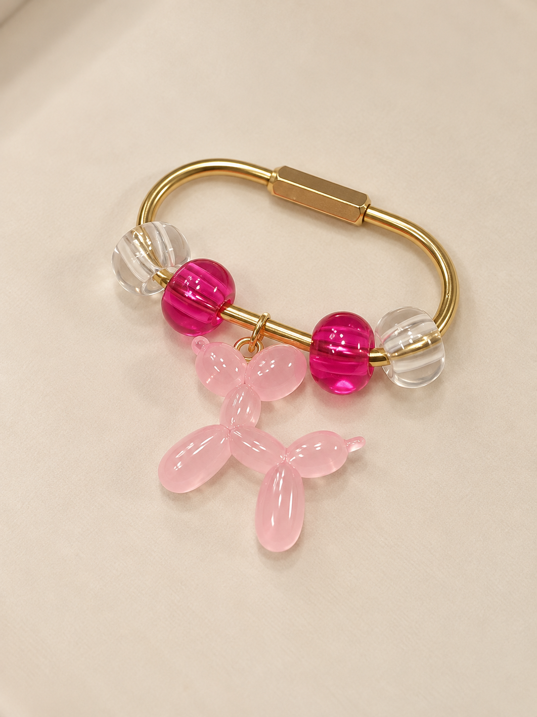 A gold-colored bracelet with pink, clear, and hot pink beads, featuring a pink balloon dog charm hanging from it, resting on a beige fabric surface.