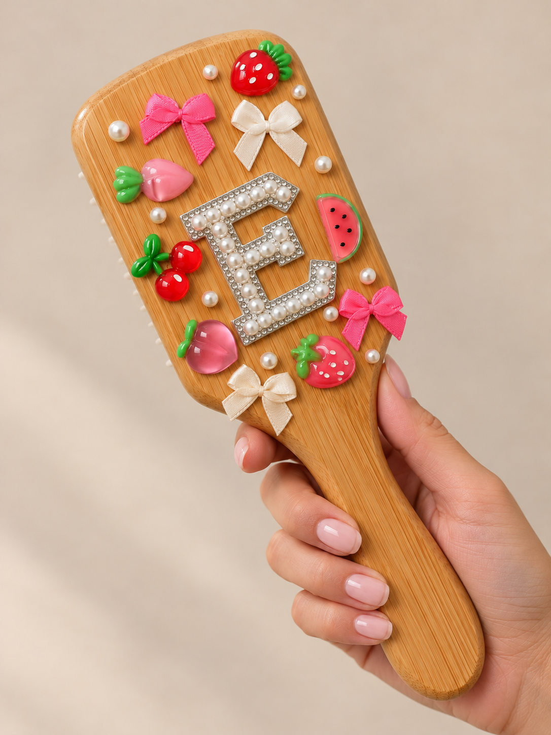 Decorative wooden hairbrush adorned with pink, red, and green plastic bows, strawberry-shaped charms, pearl beads, and a letter 'E' made of pearls and rhinestones.