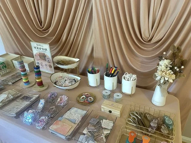 Table set up for a scrapbooking or crafting event, with various supplies including colorful washi tapes, scissors, stickers, pens, and paper, plus a sign for a scrap bar and decorative flowers as a centerpiece.