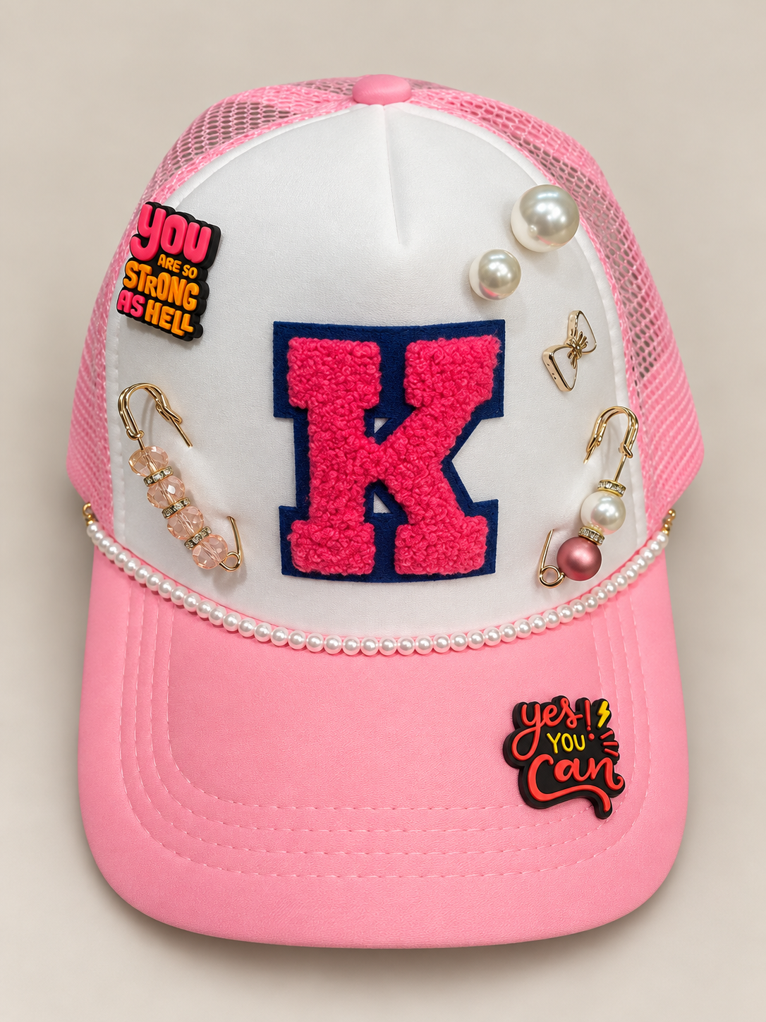 Pink and white trucker hat decorated with pins, patches, pearls, and jewelry, featuring a large pink letter K and motivational patches.