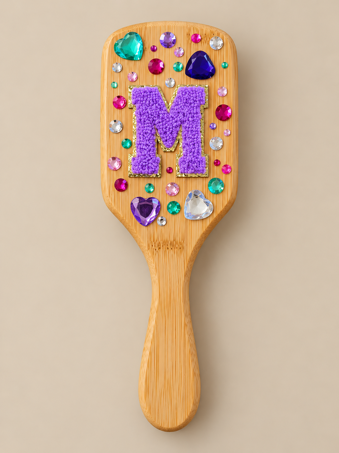 A wooden hairbrush decorated with colorful rhinestones and a large purple letter 'M' made of tiny purple flowers.