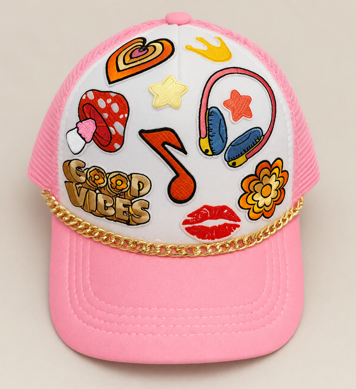 Pink baseball cap with embroidered patches of heart, star, headphones, lipstick kiss, flowers, and the phrase "Good Vibes", adorned with a gold chain detail.