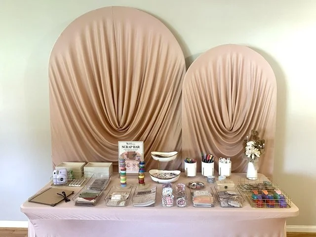 A table set up for crafting with various supplies including washi tapes, notebooks, pens, and small decorative items, against a backdrop of two large, peach-colored, draped fabric backdrops.
