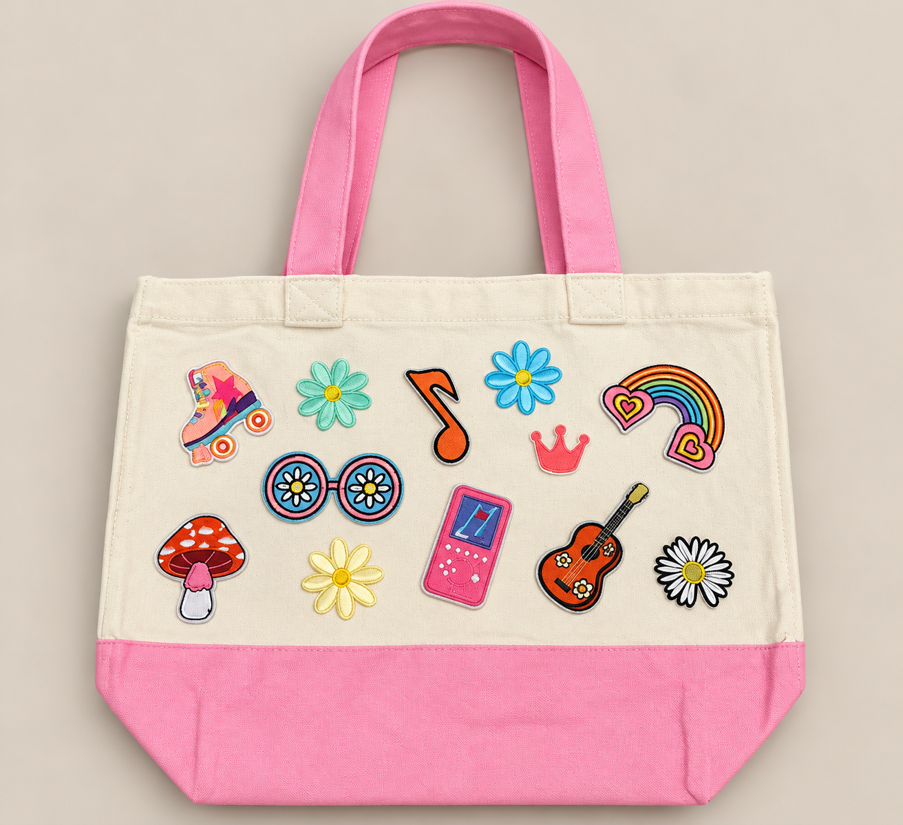 A canvas tote bag with pink handles and bottom section, decorated with colorful embroidered patches including roller skates, flowers, sunglasses, a pink music player, a guitar, a rainbow with hearts, a crown, a mushroom, and daisies.