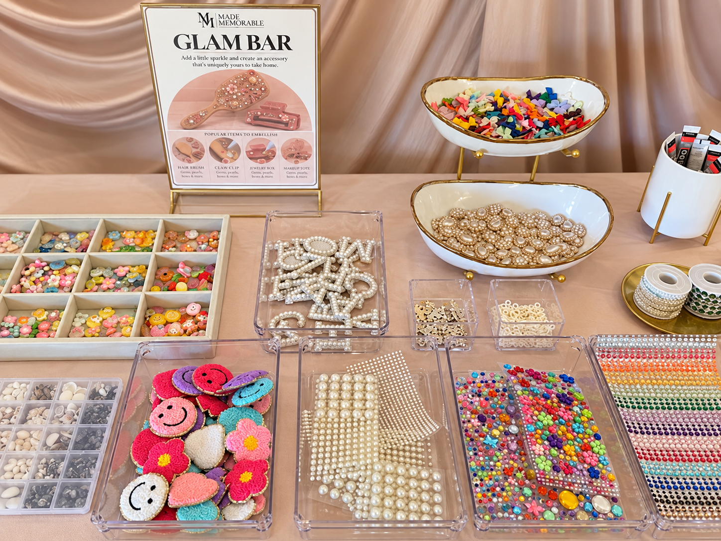 A display of colorful craft supplies including beads, buttons, smiley face patches, and rhinestones, with a sign for a glam bar activity.