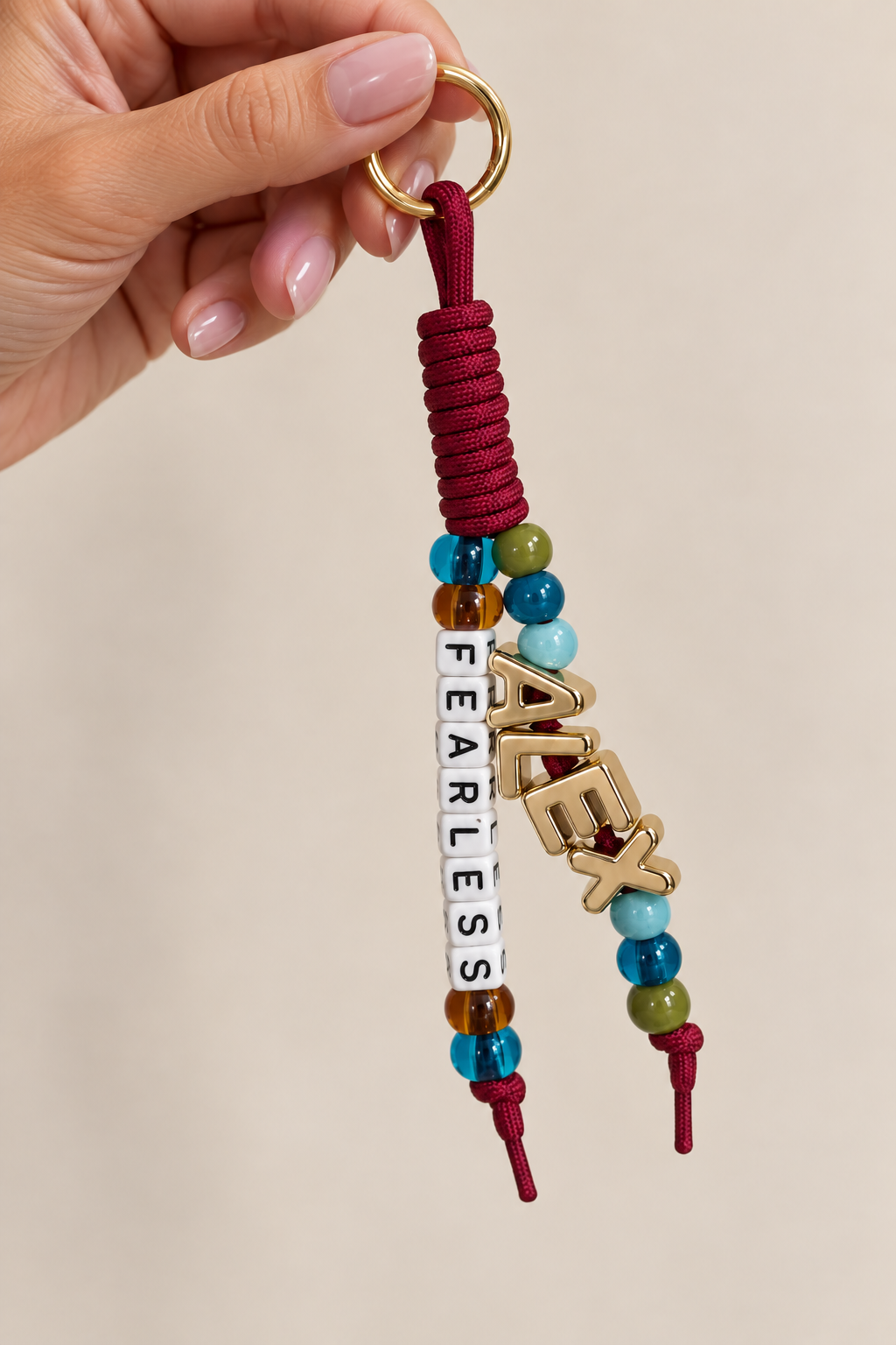 Hand holding a beaded keychain with the words 'FARLESS' and 'FLEX' on gold and white letter beads, and colorful round beads on a red cord.