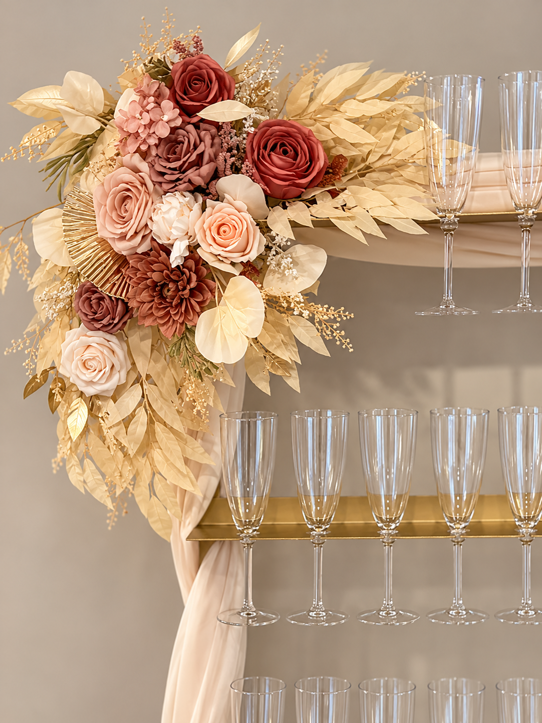 Decorative floral arrangement with pink, red, and cream roses, gold and beige leaves, and champagne flutes on a gold and beige stand.