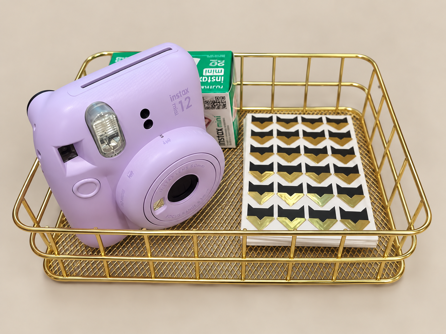 A gold wire basket containing a pastel purple instax mini 12 camera, a pack of instax mini film, and a sheet of gold and black arrow-shaped stickers on a beige surface.