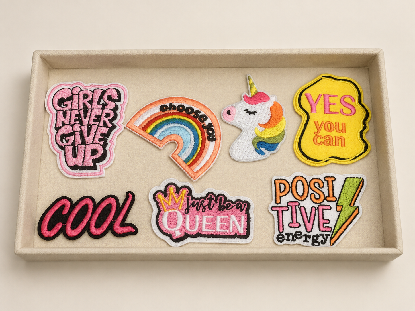 A collection of colorful embroidered patches in a box, including motivational phrases, a rainbow, a unicorn, and playful designs.
