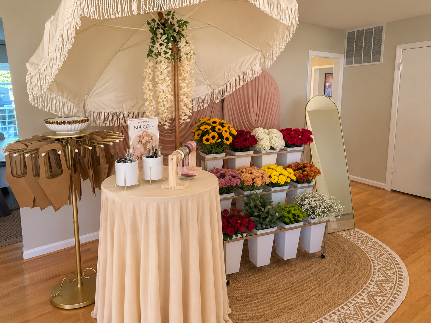A floral stand with assorted fresh flowers, a round table with a beige tablecloth holding pens and decorative items, a large umbrella with fringe, a full-length mirror, and a pink fabric backdrop behind the flowers.